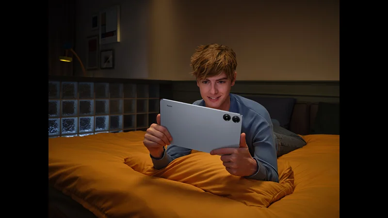 A young man is comfortably sitting on a bed, holding and using a Redmi Pad Pro 5G tablet. The tablet is in landscape orientation, and the man appears to be enjoying content on the large screen. The setting is a cozy, dimly lit room with a modern and relaxed ambiance.