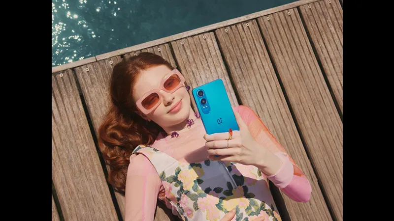 Woman with red hair wearing pink sunglasses and a floral dress, lying on a wooden deck and holding a blue OnePlus Nord CE4 Lite smartphone