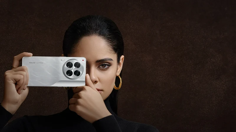 A woman holding a Tecno Camon 30 smartphone in front of her face, showcasing the phone's rear camera setup. The phone has a sleek white design and the woman is dressed in black with gold hoop earrings. The background is dark brown, highlighting the phone and the woman's features.
